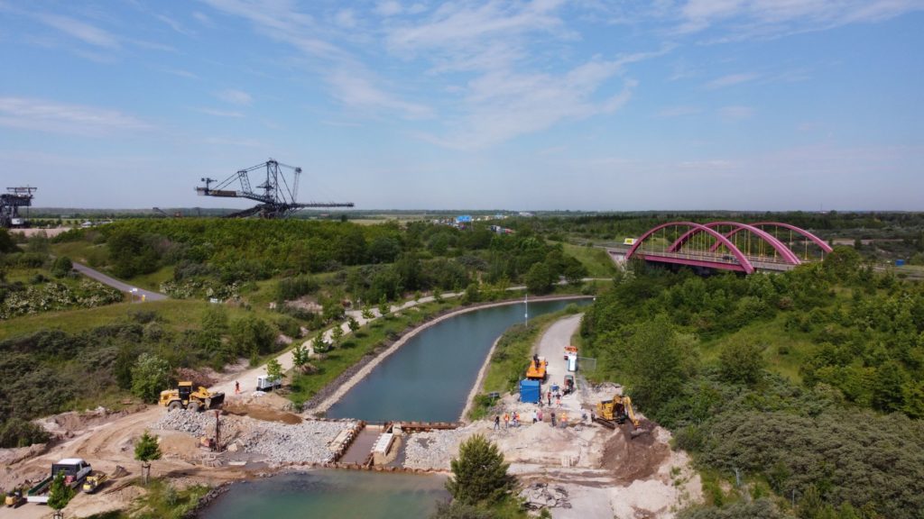 Oberes Querbauwerk ist fast fertig verfüllt, mit Wasserüberleiter in der Kanalmitte. Im Hintergrund Großgerät des Bergbau Technikpark und Brück der A,38