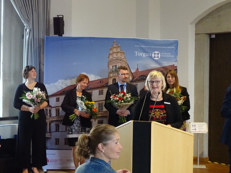 Uta Schladitz bei der Ausstellungseröffnung, am Rednerpult im Vordergrund. Im Hintergrund vier Personen mit Blumenstäußen