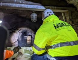 LMBV Geschäftsführer Bernd Sablotny kniet im Bereich des mit Sandsäcken errichteten Damms im Querschlag zur 1. Sohle Röhrigschacht. Er trägt eine gelbe Arbeitsschutzjacke und schaut in den Stollen, in dem Wasser steht.