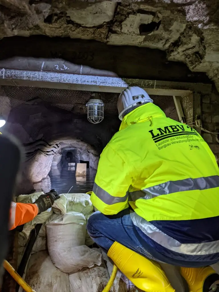 LMBV Geschäftsführer Bernd Sablotny kniet im Bereich des mit Sandsäcken errichteten Damms im Querschlag zur 1. Sohle Röhrigschacht. Er trägt eine gelbe Arbeitsschutzjacke und schaut in den Stollen, in dem Wasser steht.